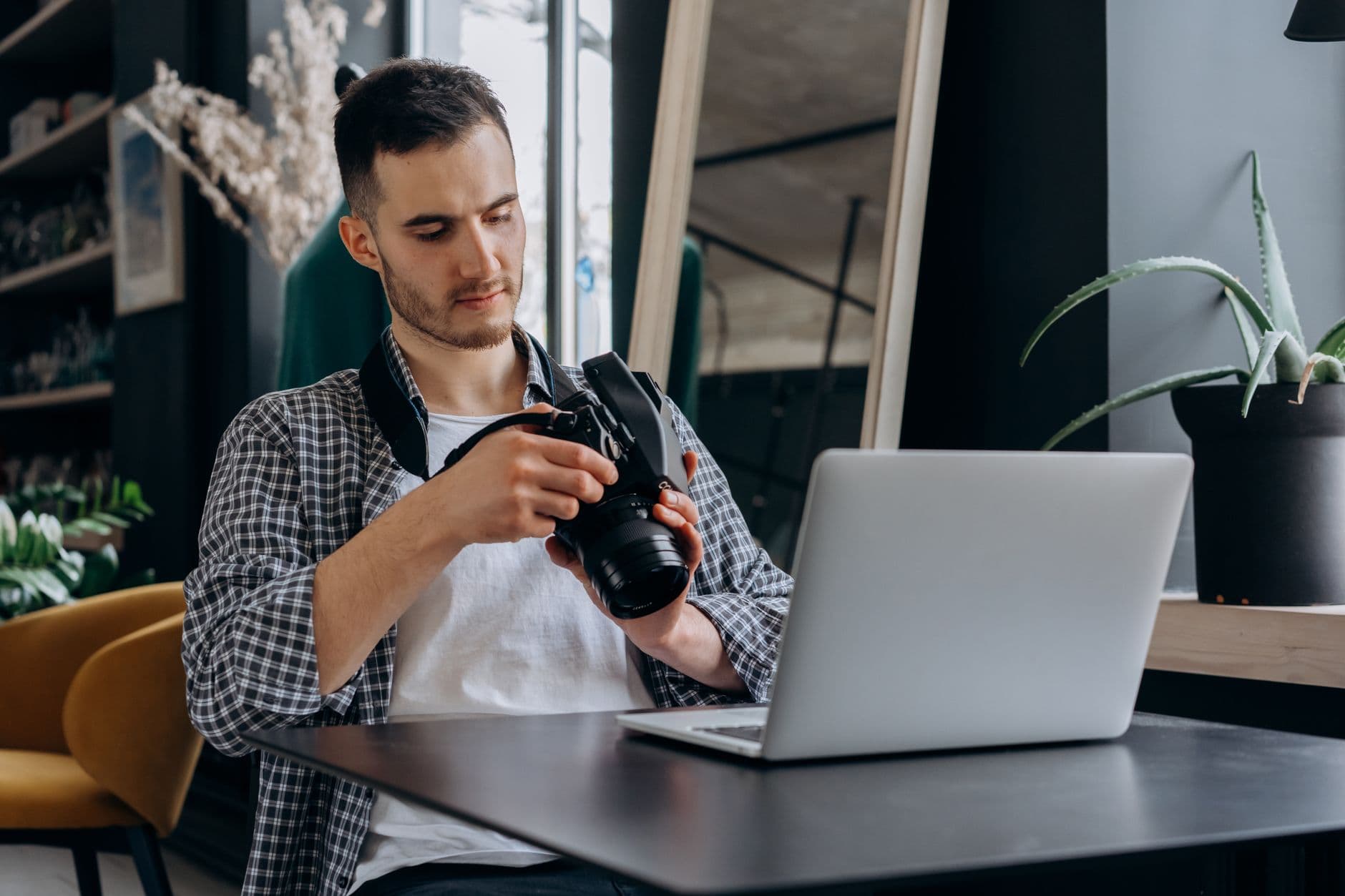 A young male photographer examines his camera while working from a laptop in a modern home office.